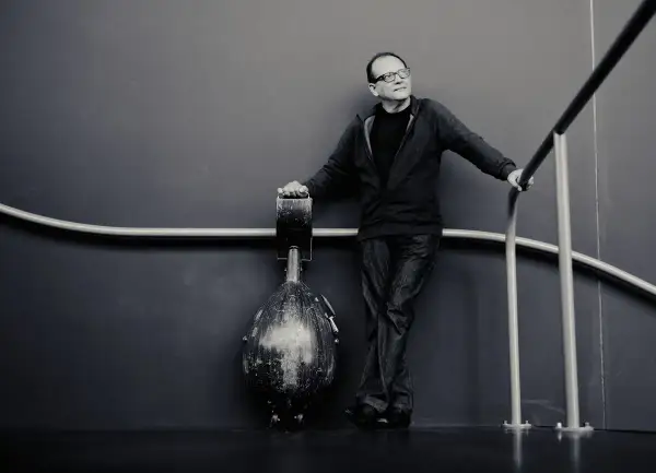 Anouar Brahem, w okularach, opiera się o poręcz, obok niego stoi instrument.