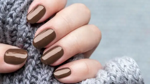 Dłonie z perfekcyjnym manicurem kombinowanym w odcieniu czekolady, otulone miękkim, szarym swetrem.