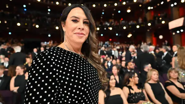 Karla Sofía Gascón, con un vestido negro de lunares blancos, sonríe en un evento con público al fondo.