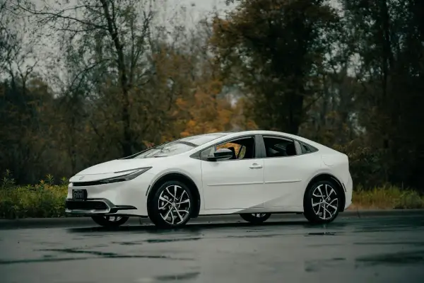 Biała Toyota Prius zaparkowana na mokrej drodze, z drzewami w tle.