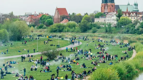 Ludzie odpoczywają na trawie w nadwarciańskiej części Poznania. W tle widać zabytkowe budynki i rzekę.