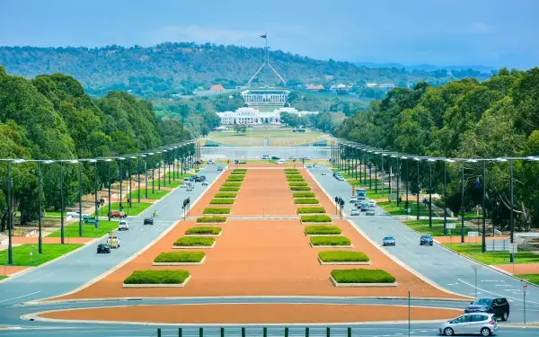 Widok na Parliament House, symbol tego, co jest stolicą Australii. Długa aleja z drzewami i samochodami prowadzi do budynku.