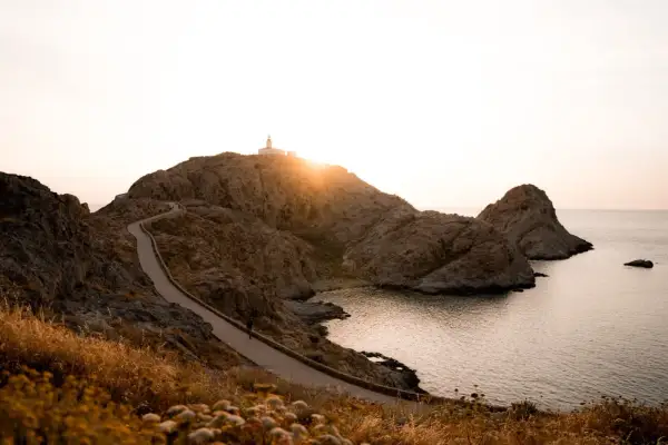 Chemin sinueux menant à un phare sur une colline rocheuse, une porte de pierre naturelle s'ouvrant sur la mer au coucher du soleil.