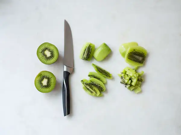 Ein Messer liegt neben zwei halbierten Kiwis. Daneben sind die Früchte in Scheiben und Würfel geschnitten, bereit zum kiwi schneiden.