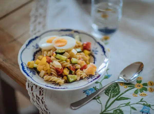 En färgglad pastasallad med ägg, grönsaker och ost, perfekt som pastasallad barn. En sked och ett glas vatten står bredvid.