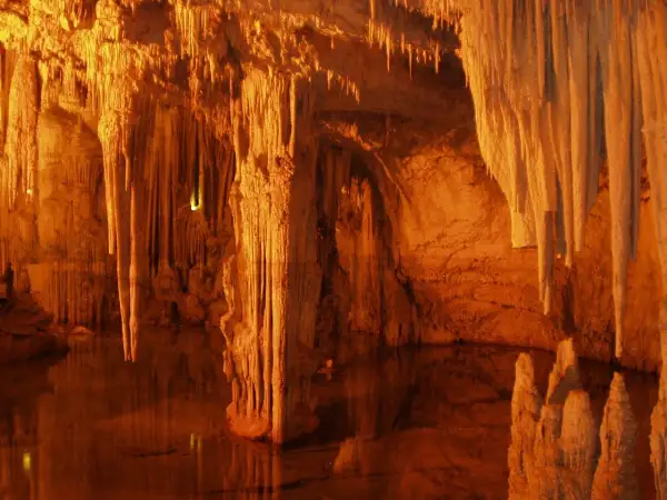 Interior de la cueva Rei Cintolo, con estalactitas y estalagmitas iluminadas en tonos cálidos, reflejándose en el agua subterránea.