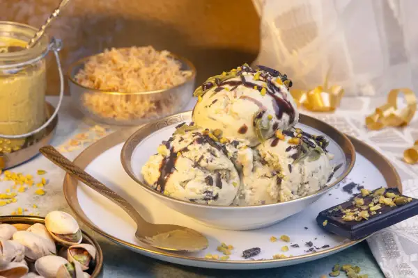 Schalen mit cremigem Pistazieneis, garniert mit Schokosauce und gehackten Pistazien. Daneben ein Stück dunkle Schokolade und eine Schale mit ganzen Pistazien.