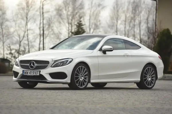 Biały Mercedes C klasa coupe z czarnym dachem, zaparkowany na brukowanej nawierzchni, z drzewami w tle.