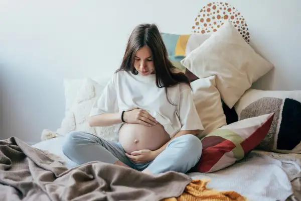 Kobieta w ciąży, otoczona poduszkami, czule dotyka brzucha. Czas oczekiwania na dziecko, być może zmagająca się z wielowodziem, ale pełna spokoju.
