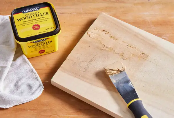 Un pot de mastic à bois Minwax est à côté d'une planche de bois où un couteau à mastic applique le produit pour reboucher bois.