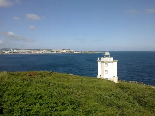 El faro de Mera se alza blanco sobre un promontorio verde, vigilando la costa y el mar azul.
