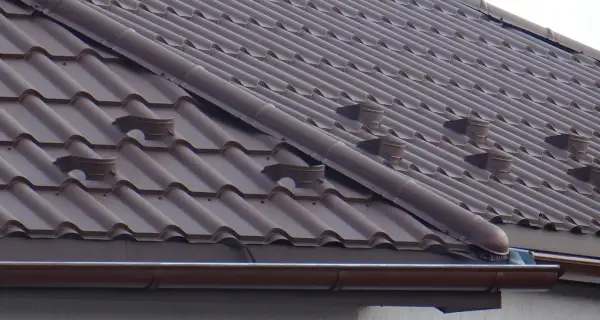 Brązowy dach z dachówki ceramicznej, widoczne elementy systemu rynnowego i wentylacyjnego. Jak zrobić zakończenie gąsiora?