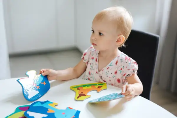 Mała dziewczynka z blond włosami skupiona na układaniu kolorowych puzzli z zwierzątkami.