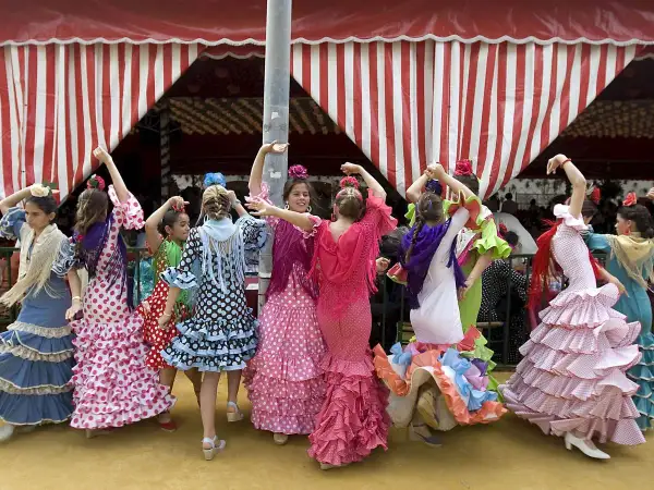 Niñas con trajes de volantes y lunares, aprendiendo a bailar sevillanas con arte bajo una carpa a rayas.