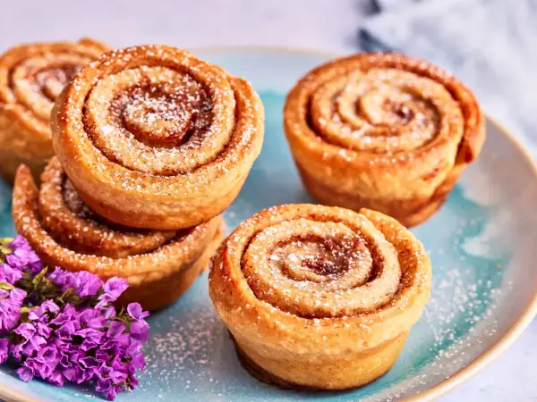 Zimtschnecken Muffins, bestäubt mit Puderzucker, liegen auf einem blauen Teller neben lila Blumen.