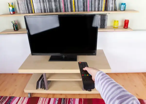 Ręka z pilotem sięga do telewizora naprzeciwko okna. Na półkach stoją płyty i kolorowe słoiki.