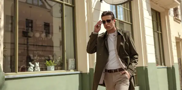 Mężczyzna w stylowym, **khaki** płaszczu i beżowych spodniach poprawia okulary, idąc ulicą.