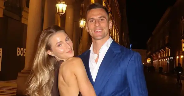 Uśmiechnięta dziewczyna z blond włosami opiera się o mężczyznę w niebieskiej marynarce. Spacerują wieczorem ulicą z latarniami.