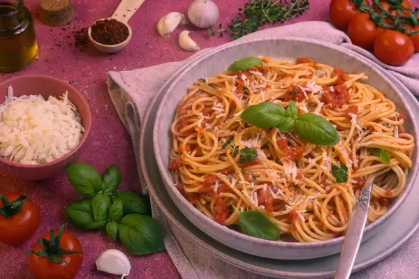 Ein Teller Spaghetti Napoli mit Tomatensauce, Basilikum und Parmesan. Daneben frische Tomaten, Knoblauch und Kräuter.