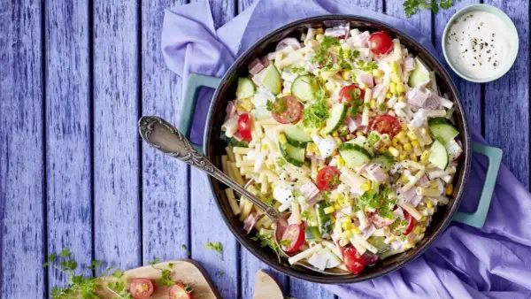 Ein köstlicher nudelsalat mit schinken, garniert mit Kirschtomaten, Gurkenscheiben und Mais. Perfekt für ein Sommerfest!