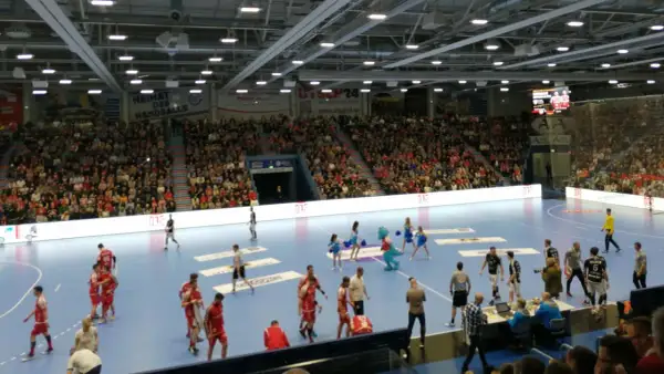 Handballspiel in der Gummersbach Schwalbe Arena. Zuschauer füllen die Ränge, Spieler auf dem Feld, eine Maskottchen-Figur und Cheerleader sind zu sehen.