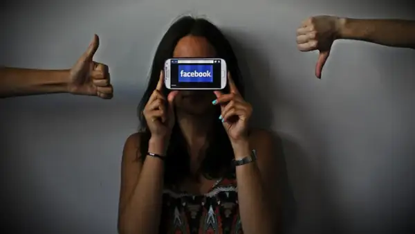 Una persona oculta su rostro tras un móvil con Facebook. A los lados, pulgares arriba y abajo, reflejando el impacto de las redes sociales y la autoestima.