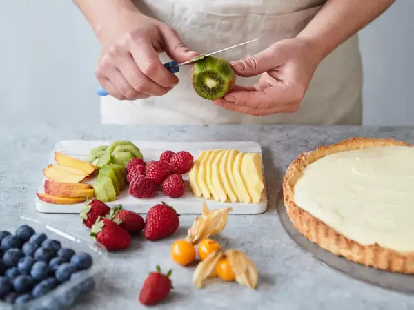 Hände schälen Kiwi für ein fruchtiges Tarte Rezept. Daneben liegen Beeren, Pfirsichspalten und Apfelscheiben.