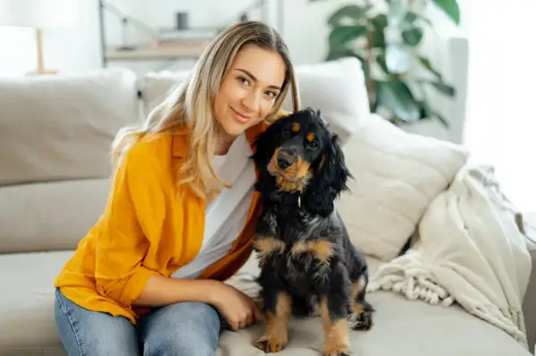 Uśmiechnięta blondynka i jej piesek siedzą na kanapie. Piesek patrzy w obiektyw, jakby chciał powiedzieć: "dlaczego pies chodzi za mną krok w krok?".