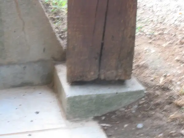 Un poteau bois pourri à la base repose sur un socle en béton. La dégradation du bois est visible.