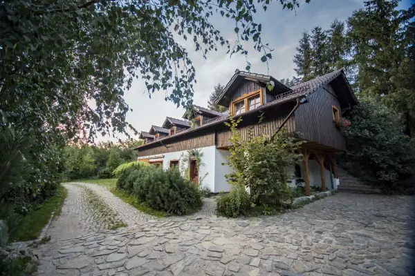 Urokliwa stajnia pod świerkami, otoczona zielenią, z kamienistą ścieżką prowadzącą do wejścia.