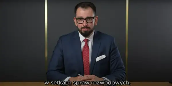 Mężczyzna w garniturze i okularach siedzi przy biurku, trzymając długopis.