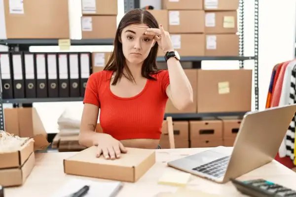 Mujer frustrada frente a un portátil, rodeada de cajas y material de oficina, lidiando con la gestión de stock.