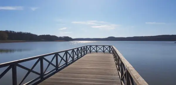 Holzsteg am Schmachter See lädt zum Spaziergang auf dem Rundweg ein. Sonnenschein glitzert auf dem Wasser, bewaldete Ufer im Hintergrund.