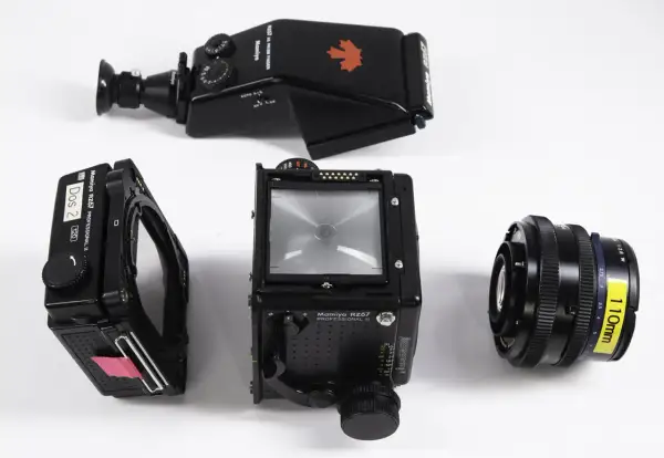 A Mamiya RZ67 camera body, film back, prism finder, and 110mm lens are laid out on a white surface.