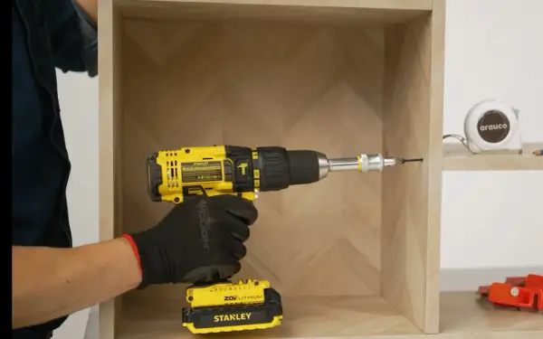 Mano con guante negro usa taladro amarillo para unir dos muebles. Cinta métrica y herramientas al fondo.