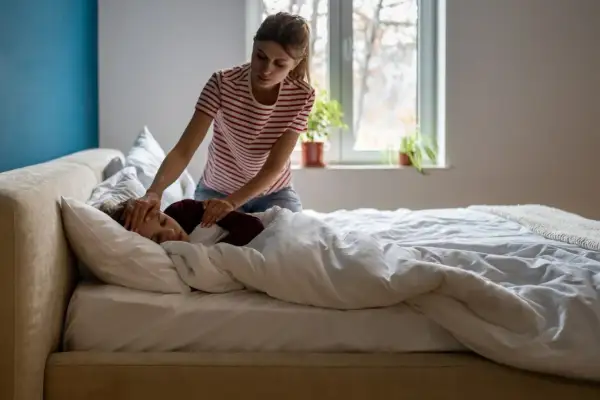 Kobieta sprawdza czoło śpiącego dziecka, które leży w łóżku pod kołdrą. Wygląda na to, że dziecko ma przeziębienie.