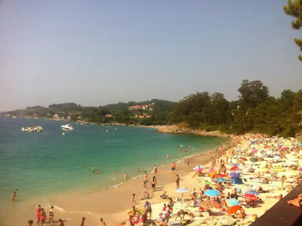Gente disfrutando del sol y el mar en la playa areacova, con barcos en el agua y colinas verdes al fondo.