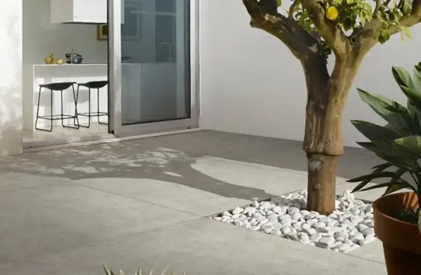 Un moderno patio con diversi tipi di pavimentazione esterna: lastre in gres grigio e un'area con ciottoli bianchi attorno a un albero.