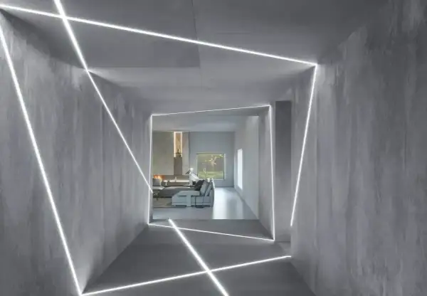Nowoczesny korytarz z betonowymi ścianami i sufitem, ozdobiony geometrycznymi liniami światła LED. W tle widać salon z kominkiem i meblami. To inspiracja, jak zamontować oświetlenie LED w witrynie.