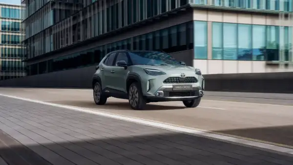 En grön Toyota Yaris Cross hybrid kör på en stadsgata. Bilen är i fokus framför moderna byggnader.