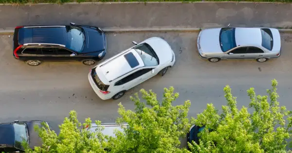 Biały SUV wykonuje parkowanie równoległe przodem między czarnym SUV-em a srebrnym sedanem.