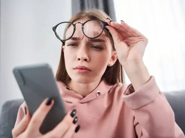 Zmarszczona kobieta w okularach trzyma telefon, szukając domowych sposobów na mroczki przed oczami.