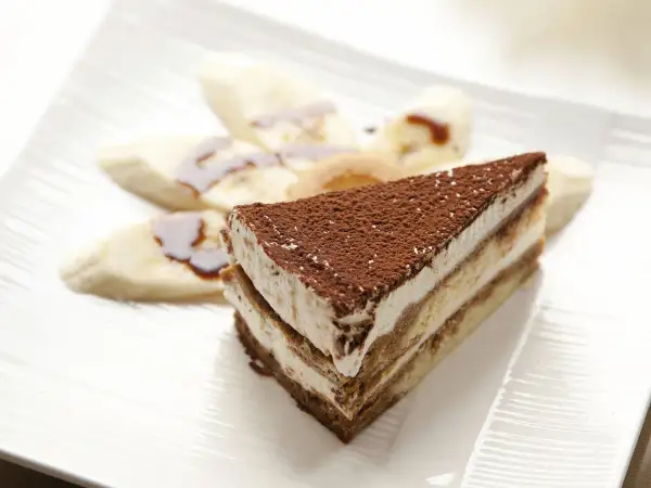 Tranche de tiramisu banane, saupoudrée de cacao, accompagnée de rondelles de banane nappées de sauce. Un délice !