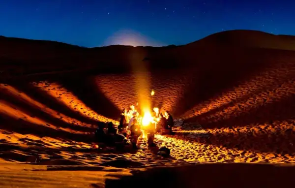 Un groupe se réchauffe autour d'un feu de camp dans le désert du Sahara la nuit. La température chute, mais la chaleur du feu et la beauté du ciel étoilé créent une atmosphère magique.