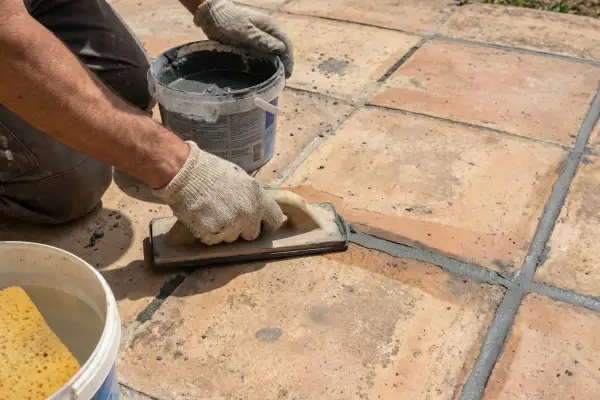 Application d'enduit pour joint carrelage extérieur. Un artisan, ganté, lisse le mortier gris avec une taloche sur des carreaux de terre cuite.