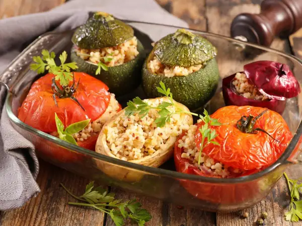 Un plat de petits farcis gastronomiques : tomates, courgettes rondes et oignons rouges farcis de grains et herbes, dans un plat en verre.