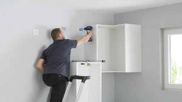 Hombre en escalera usa taladro para fijar soportes en pared, parte de como colocar kit para colgar armarios.