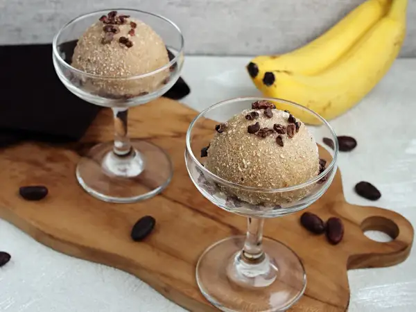 Zwei Gläser mit cremigem Apfel Zimt Eis, garniert mit Kakaonibs. Bananen im Hintergrund.