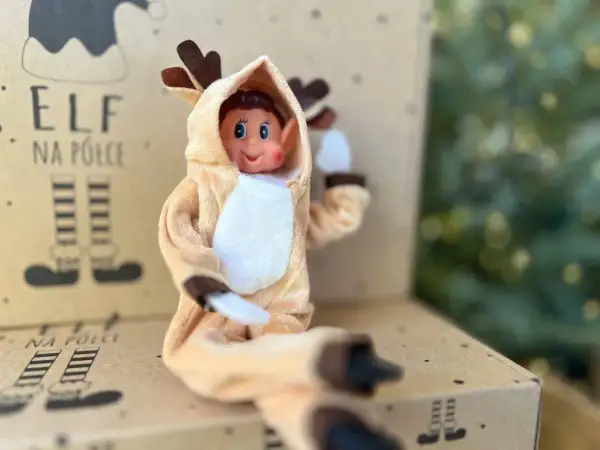 Uroczy elf w stroju renifera z uszkami i brązowymi rogami siedzi na kartonie z napisem "Elf na półce".