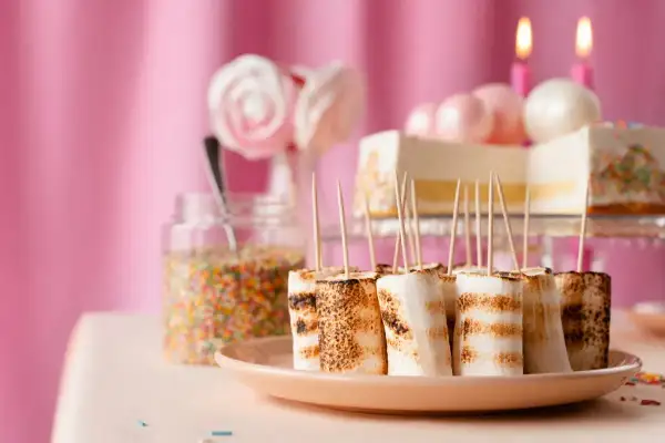 Grillade marshmallows på spett, perfekt fika till barnkalas. En tårta och strössel i bakgrunden.